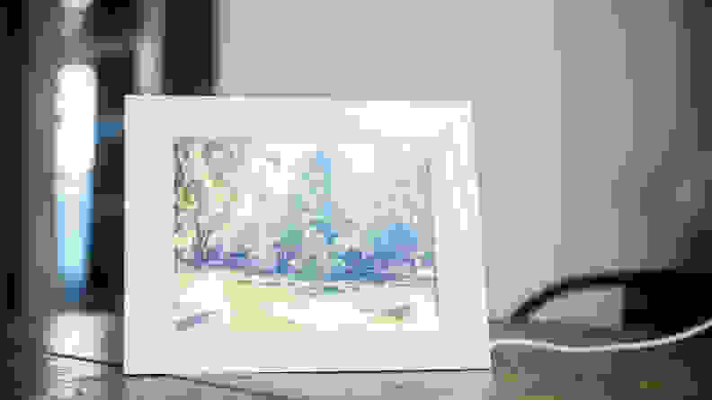 A white Aura digital picture frame displays an image of a brick building surrounded by trees.