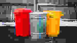 A red Rubbermaid 32 gallon outdoor trash can, Behrens galvanized steel 31 gallon trash can, and a yellow Suncast trash can sitting outside in front of a fence.