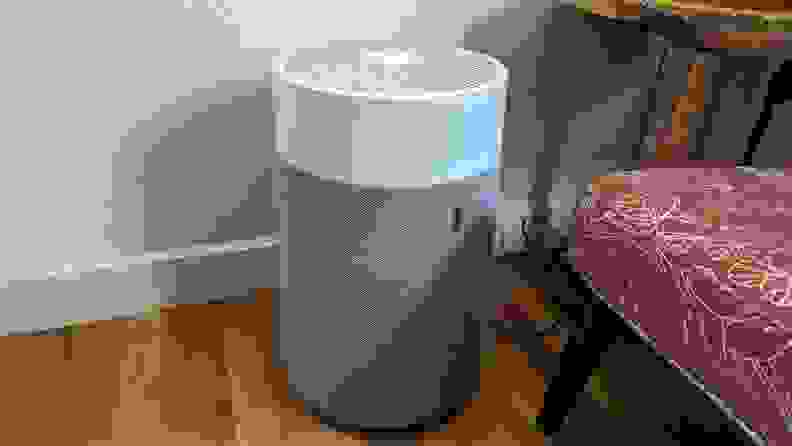 Gray and white Blueair Blue Pure 311i Max purifier sitting on top of wooden floor boards next to padded chair in front of white wall.