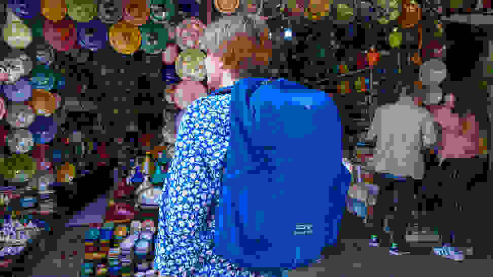 A woman wearing a blue Osprey travel backpack stands outside a pottery vendor in a Morocco market.