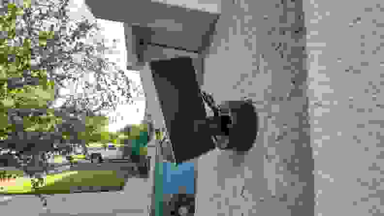 The Eve Outdoor Cam smart floodlight hands on the side of a garage wall on the exterior of a home with a neighborhood view in the background