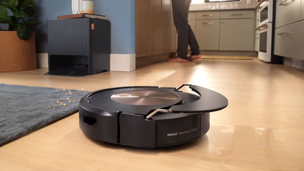 A black, round robot vacuum on a light wooden floor next to a gray rug, with a person standing in the background