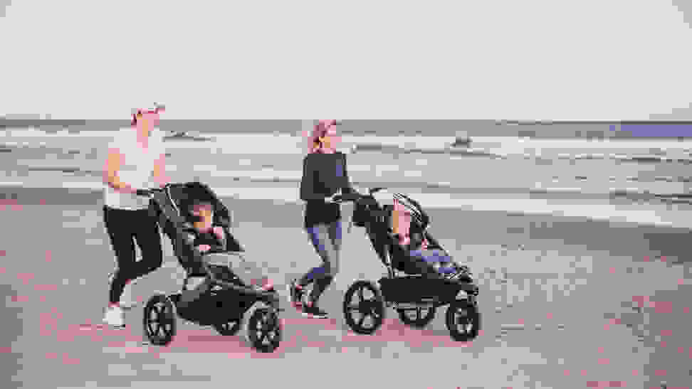 Two women jogging on a beach pushing kids in jogging strollers