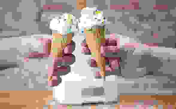 Two hands holding ice cream cones with vanilla ice cream and rainbow sprinkles cheers in front of an ice cream maker on a wood counter in front of an exposed brick wall.