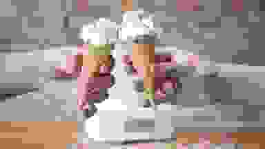 Two hands holding ice cream cones with vanilla ice cream and rainbow sprinkles cheers in front of an ice cream maker on a wood counter in front of an exposed brick wall.