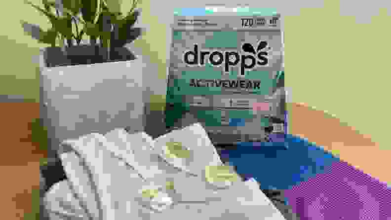 A box of Dropps detergent sits on a table with some clothes and a plant