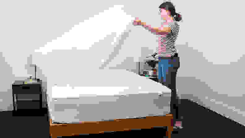 a person spreads a sheet above a mattress with a mattress protector on it