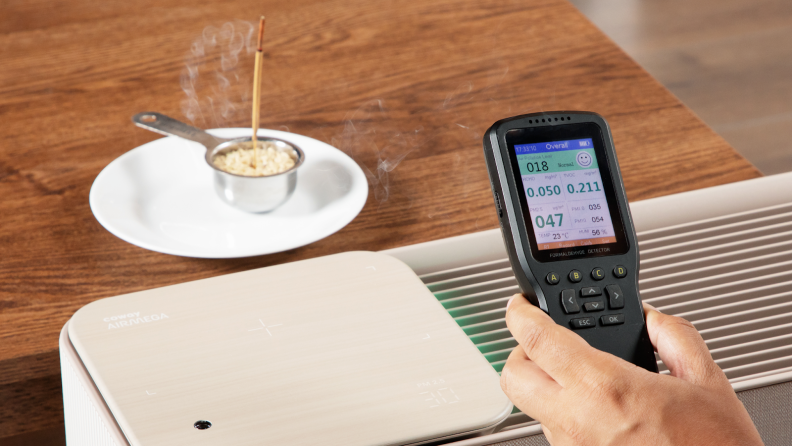 Incense burns, sticking up from a measuring cup of rice, while above a hand holds an air monitor.