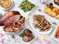 A buffet of Easter dinner options spread on a marble counter: ham, lamb roast, lamb chops, quiche, green beans, carrots, a cheese board, and hot cross buns, surrounded by bits of colorful Easter candy.