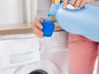 Person pouring liquid laundry detergent in cup in front of washing machine.