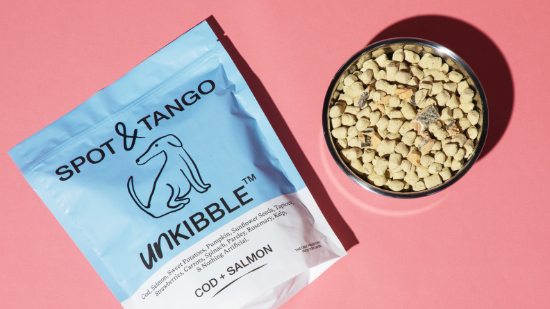 A bag of spot and tango dog food next to a silver bowl of dog food on a pink background