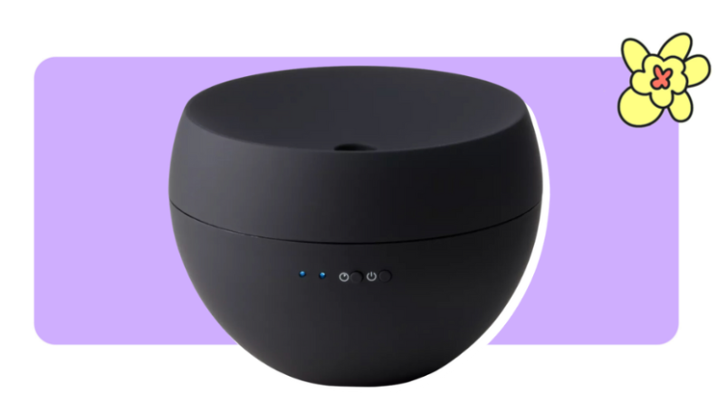 A black Stadler Form Aromatherapy Oil Diffuser in front of purple background with a yellow cartoon flower in the corner.