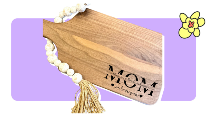 A wooden cutting board with the words carved into the bottom right with a small string of wooden beads underneath in front of purple background with a yellow cartoon flower in the corner.