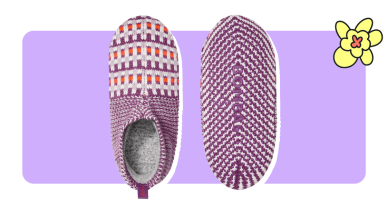 A pair of knitted Bombas Gripper Slippers in Sweet Violet color in front of purple background with a yellow flower in the corner.